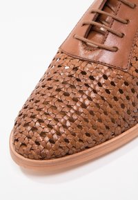 Chaussure en cuir marron avec un motif tissé sur l'avant-pied et une fermeture à lacets, posée sur fond blanc.