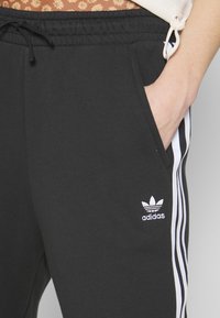 Černé tepláky vyrobené z kombinace bavlny, s bílým logem Adidas, bočními kapsami a kontrastními bílými pruhy po stranách.