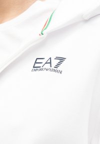 Λευκή φούτερ με κουκούλα με κεντημένο λογότυπο "EA7 Emporio Armani" σε ναυτικό μπλε. Η κουκούλα έχει λεπτομέρεια με πολύχρωμη ρίγα σε κόκκινο, λευκό και πράσινο.