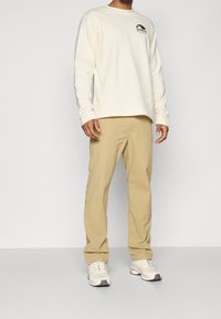 Vaalean kermanvärinen pitkähihainen paita, jossa pieni logo rinnassa, yhdistetty beigeihin housuihin ja valkoisiin urheilukengiin. Tasainen kangas ja rento istuvuus.