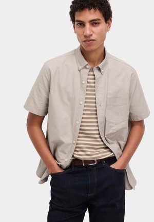Ung mand iført en beige kortærmet skjorte med knapper over en beige og hvidstribet t-shirt, med mørke jeans og hænderne i lommerne.