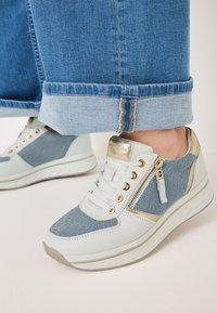 Sneakers bianchi e in denim con design con lacci, dettagli dorati, cerniera laterale e suola testurizzata. Jeans blu risvoltati che si fermano sopra le sneakers.