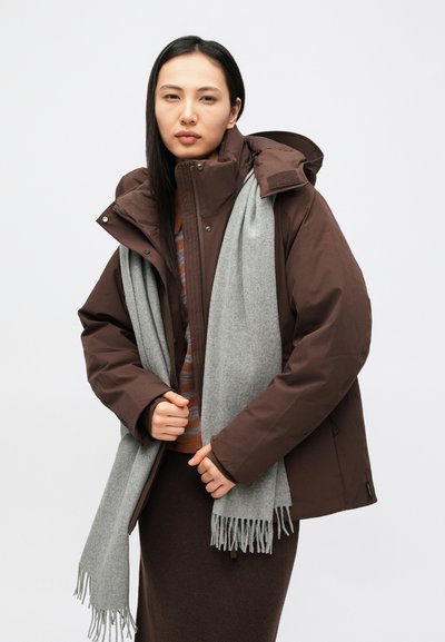 Brązowa ocieplana kurtka z wysokim kołnierzem i kapturem, w połączeniu z jasnoszarym szalikiem z frędzlami. Model stoi na jednolitym tle.