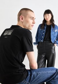 Camiseta negra con texto gráfico blanco en la parte de atrás, jeans de mezclilla azules, y una mujer con una blusa negra y una chaqueta de mezclilla azul al fondo.