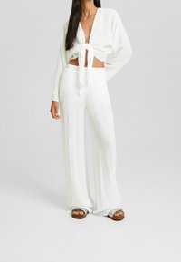 Blusa bianca corta con nodo frontale e maniche lunghe, abbinata a pantaloni bianchi larghi e morbidi. Il tessuto testurizzato aggiunge un sottile dettaglio.