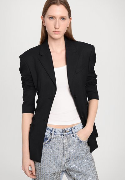 Black tailored blazer with shoulder pads, single-breasted design, and three-quarter sleeves. Worn over a white top and embellished denim jeans.