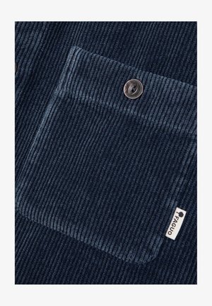 Marineblauwe corduroy stof met verticale ribben, een enkele borstzak met een metalen knoop en een klein wit merklabel.