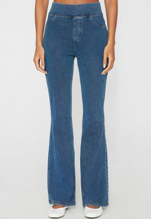Donna che indossa jeans a campana in denim blu a vita alta con tasche frontali e scarpe basse bianche, in piedi su uno sfondo semplice.