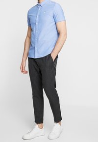 Camicetta azzurro chiaro a maniche corte con colletto e piccolo emblema verde, abbinata a pantaloni scuri a righe e sneakers bianche.