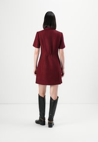 Donna in piedi di spalle con un vestito rosso a maniche corte e lunghezza al ginocchio, abbinato a stivali in pelle nera al ginocchio con dettagli in catena dorata.