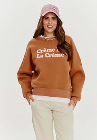 Hnědá mikina s růžovým textem "Crème de La Crème", volný střih, žebrované manžety, nošená přes bílou košili, doplněná růžovou kšiltovkou.