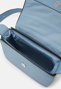 Světle modrá obdélníková crossbody taška otevřená, aby ukázala zipovou vnitřní kapsu a magnetické zapínání na bílém pozadí.
