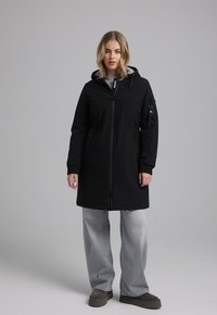 Zwarte knie-lange jas met een capuchon, ritsluiting, zijzakken en verstelbare trekkoorden in de taille. Combinatie met grijze wijde jeans.