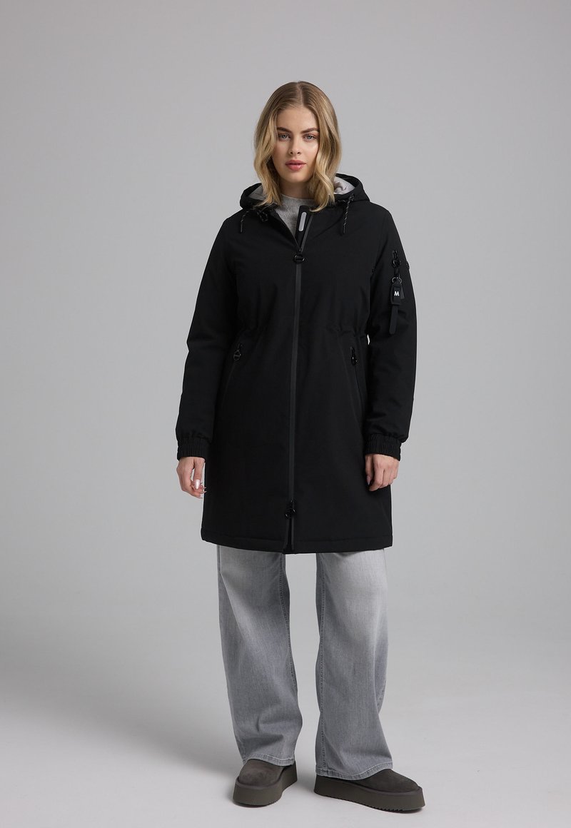 Zwarte knie-lange jas met een capuchon, ritsluiting, zijzakken en verstelbare trekkoorden in de taille. Combinatie met grijze wijde jeans.