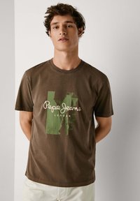 Camiseta de algodón marrón con un diseño gráfico en verde que incluye el texto "Pepe Jeans London". Mangas cortas y cuello redondo.