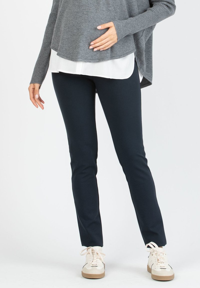 Persona con pantalones negros ajustados, zapatillas blancas, suéter gris sobre camisa blanca, con una mano apoyada en el estómago.