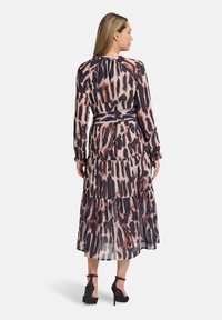 Fließendes, gestuftes Kleid aus transparenter Stoff mit einem braun-schwarzen abstrakten Muster, langen Ärmeln und einem passenden Gürtel. Schuhe mit Absatz vervollständigen den Look.