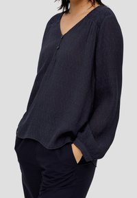 Marinblus med V-ringning, med subtil textur och samlade axeldetaljer. Avslutad med en knappstängning vid halsen.