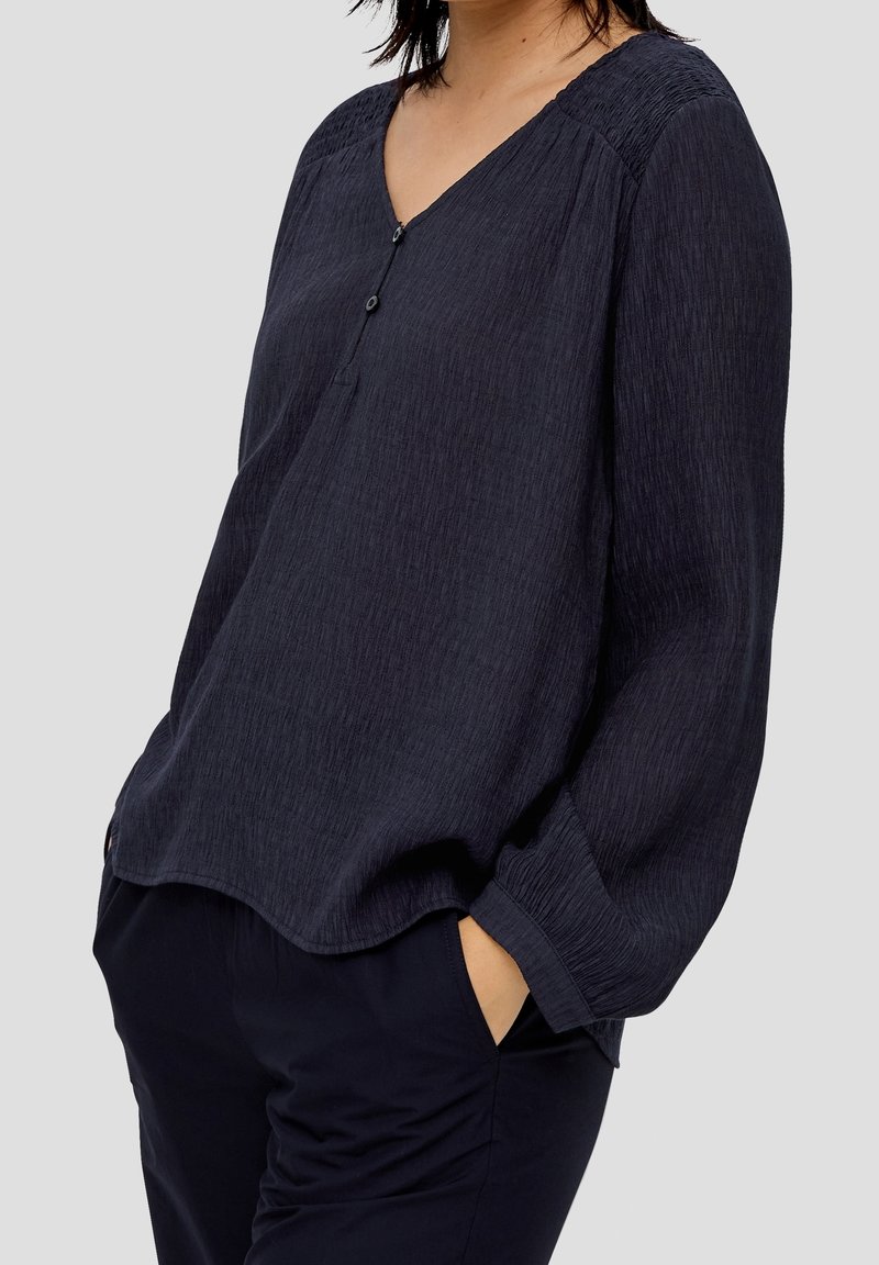 Marinblus med V-ringning, med subtil textur och samlade axeldetaljer. Avslutad med en knappstängning vid halsen.