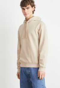 Bež hoodie sa vezicom, mekani materijal, kengurska džep, rebrasti rukavi i rub, uparen sa svijetloplavim farmerkama u minimalističkom okruženju.