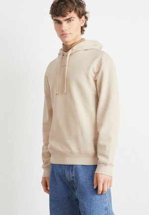 Sweat à capuche beige avec cordon de serrage, en tissu doux, poche kangourou, manches et bas côtelés, accompagné de jeans bleu clair dans un cadre minimaliste.