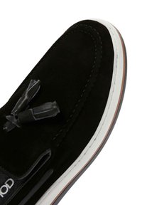 Black suede loafer with a rounded toe, white rubber sole, and contrasting stitching. Features dual black tassels on the front.