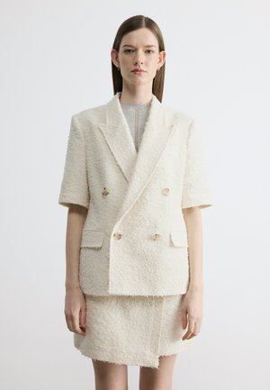 Kobieta o prostych brązowych włosach, ubrana w teksturowaną, dwurzędową, kremową marynarkę z krótkimi rękawami oraz dołączoną spódnicę, stojąca na białym tle.