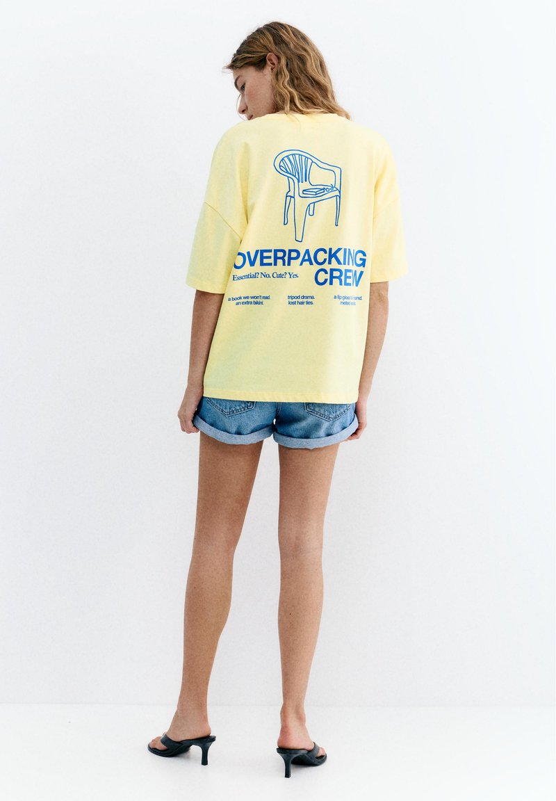 Femme en t-shirt jaune oversize avec un graphisme de chaise bleue et le texte « Overpacking Crew », short en jean et sandales à talons noires, debout face au mur.