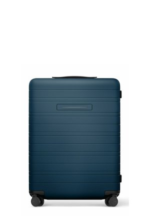 Valise rigide bleu foncé avec des rainures horizontales, poignée supérieure, quatre roues pivotantes noires et une zone rectangulaire encastrée pour la plaque nominative.