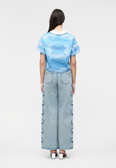 Γυναίκα με μακριά σκούρα μαλλιά φοράει μπλε με σχέδια κοντομάνικη μπλούζα και φαρδιά ανοιχτόχρωμα τζιν με λεπτομέρειες στα πλάγια και σανδάλια με ψηλό τακούνι.