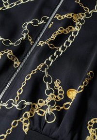 Tissu noir imprimé de chaînes dorées et beiges, avec une fermeture éclair centrale argentée et un détail de ceinture élastique noire.