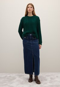Woman wearing a dark green knit sweater, long dark blue denim skirt with front slit, and dark brown ankle boots, standing against a plain wall.