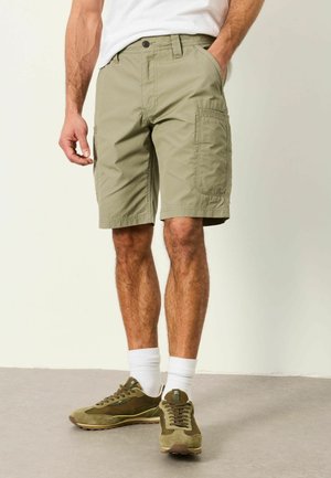 Mann trägt beige Cargo-Shorts, weiße Socken und olivgrüne Sneakers, steht mit einer Hand in der Tasche vor einfarbigem Hintergrund.