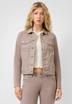 Junge Frau mit welligen blonden Haaren, die eine hellbraune, geknöpfte Jeansjacke, passende Hosen und ein weißes, gemustertes, bauchfreies Oberteil trägt.