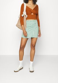 Top corto lavorato a maglia marrone con dettaglio a torsione, abbinato a una mini gonna a quadri verde e bianca. Sneakers bianche e una borsa a tracolla beige completano il look.