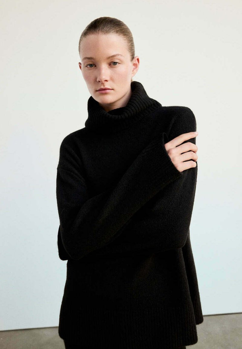 Jeune femme aux cheveux coiffés en arrière portant un pull à col roulé noir oversize, bras croisés, sur un fond clair uni.