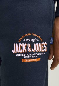 Sötétkék joggingnadrág kiemelkedő fehér és narancssárga logóval, amelyen a "JACK & JONES" felirat található, valamint egy kis kék szövetcímke az oldalon.