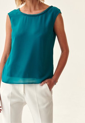 Blouse sans manches en teal avec un col tressé texturé, associée à un pantalon blanc sur mesure. Tissu lisse avec une coupe décontractée.