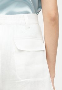 Κοντινό πλάνο λευκών παντελονιών με ψηλή μέση και ελαστική μέση με τσέπη με καπάκι, που φοριούνται με ανοιχτό μπλε σατέν τοπ, δείχνοντας το χέρι και το πλευρικό κορμό.