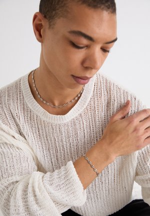 Zilveren ketting en armband op een persoon die een textuurloos, losvallend wit trui draagt. De trui heeft een brede halslijn en ribbeldetails.