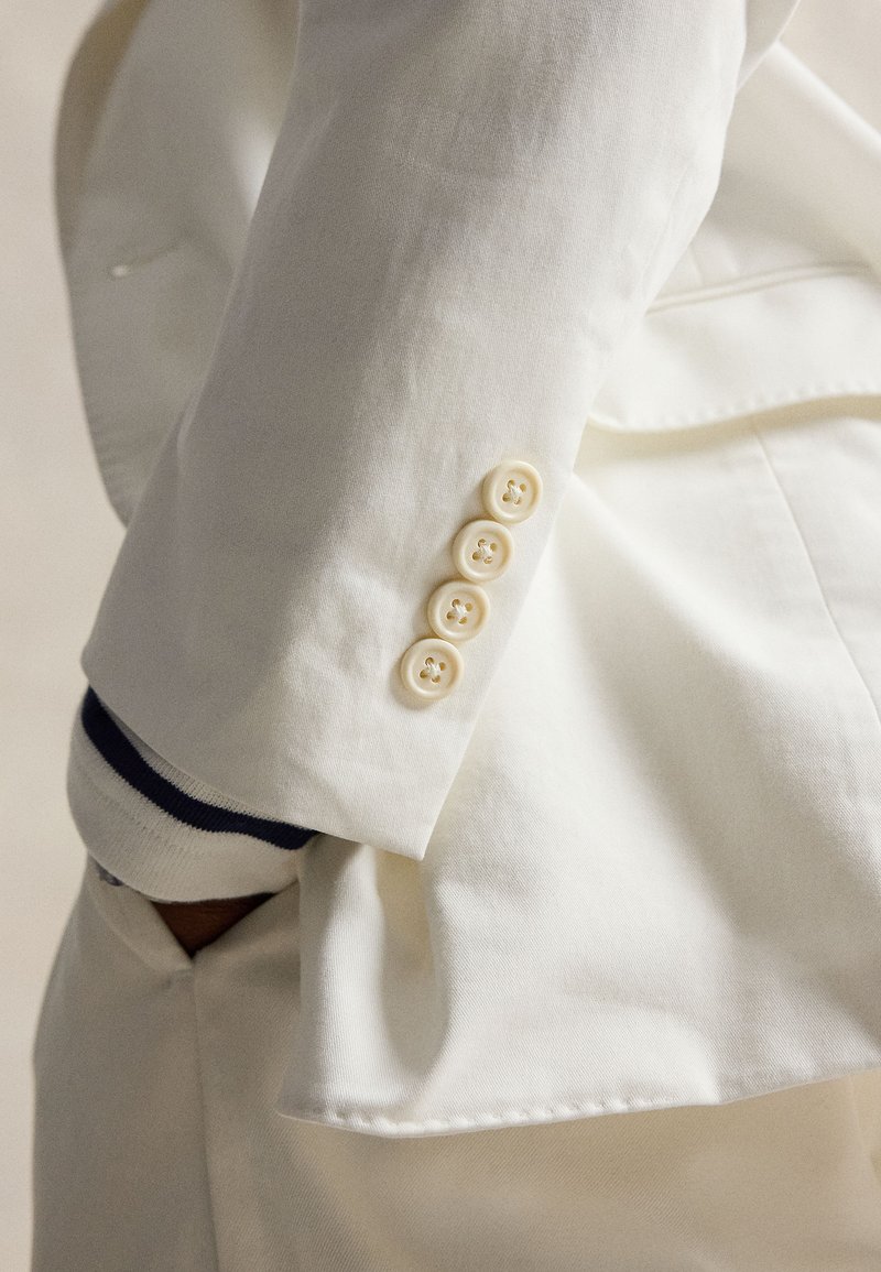 Manche de blazer blanche avec quatre boutons ivoire sur un poignet rayé, main glissée dans la poche du pantalon blanc assorti.