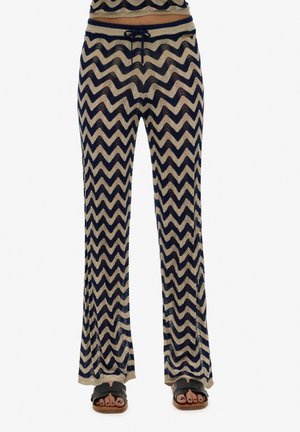 Knit pants with a chevron pattern in navy and beige, finished with a drawstring waistband and flared legs, paired with slide sandals.