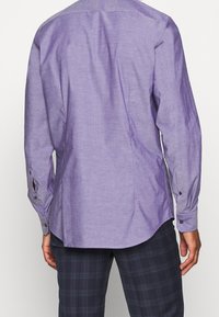 Fialová košile s dlouhým rukávem z hladké látky, s manžetami na knoflíky a zakřiveným spodním okrajem. Na zadní straně je sedlo a boční švy.