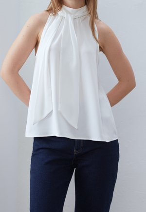 Femme portant un haut blanc sans manches avec un détail de nœud au col montant et un jean bleu foncé, mains reposant dans le dos.