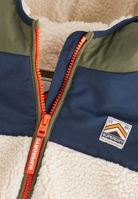 Chaqueta de forro polar con paneles azul marino y verde oliva, cremallera naranja y una parche de tela con un logo de montaña. Interior suave y texturizado.