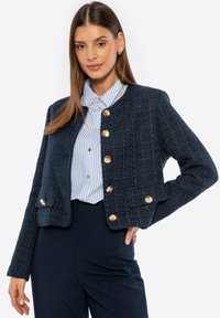 Námořnický tvídový blazer s texturovaným povrchem, zlatými knoflíky a zkráceným designem. Nosený přes světlomodrou pruhovanou košili.