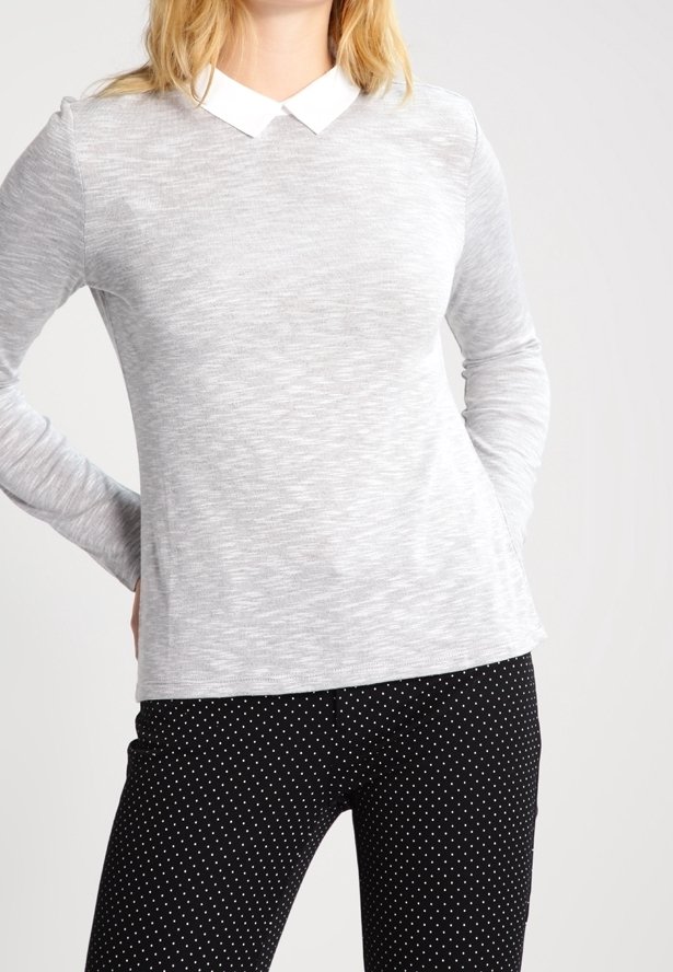 Femme portant une chemise gris clair à manches longues avec col blanc et un pantalon noir à petits pois blancs, debout les mains dans le dos.