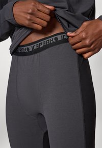 Šedé sportovní legíny s černým elastickým pasem, na kterém se opakuje logo. Materiál vypadá hladce s texturovaným švem.