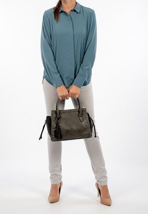 Frau in einem türkisfarbenen Hemd mit Knöpfen und hellen Hosen, die eine dunkle Ledertasche mit Quasten hält, beige High Heels trägt und vor weißem Hintergrund steht.