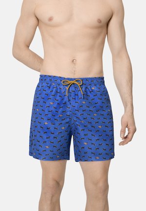 Hombre con pantalones cortos de baño azules con pequeños patrones de perros negros y marrones, y un cordón amarillo, mostrado desde el pecho hasta la mitad del muslo.